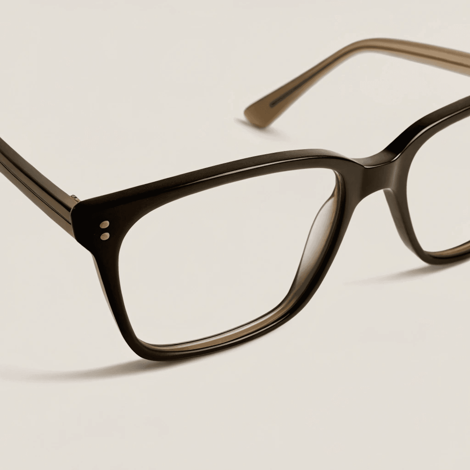 Close detail of dark eyeglass frames with two small rivets on a warm ivory background.